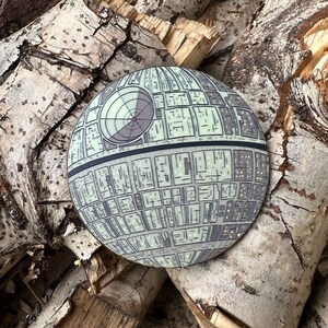 May include: A round, gray and white Death Star pin with a detailed design. The pin is sitting on a piece of wood.