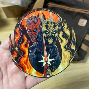 Pode incluir: Um coaster de madeira circular com uma imagem colorida de dois personagens de Star Wars, Darth Maul e Darth Sidious, com chamas e um sabre de luz no fundo.