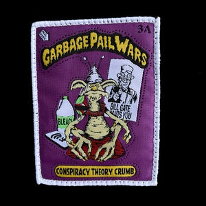May include: A purple and white embroidered patch with the text "Garbage Pail Wars" in yellow letters. The patch features a cartoon alien holding a bottle of bleach and a sign that says "Bill Gate Wants You". The bottom of the patch says "Conspiracy Theory Crumb".