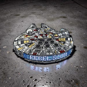 May include: A detailed patch featuring the Millennium Falcon starship from Star Wars. The patch is white, gray, yellow, and red, with the text "REBEL SCUM" in blue. The patch is on a gray surface.