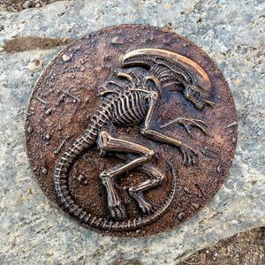 May include: A circular wall plaque featuring a relief sculpture of a Xenomorph creature in a bronze-toned finish. The alien figure is depicted in a crouched, skeletal pose with an elongated head, ribbed torso, and a long, curved tail.