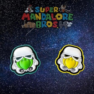 May include: Two white and black cartoon stormtrooper helmet patches with green and yellow faces. The patches are on a black background with white stars.