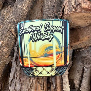 May include: A glass of whiskey with the text "Emotional Support Whiskey" on it. The glass is filled with a yellow liquid and has a checkered pattern on the bottom.