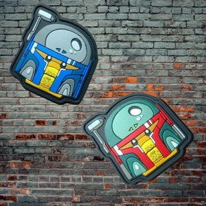 May include: Two cartoon-style patches featuring a blue and green Boba Fett character with a yellow helmet and a red stripe. The patches are made of a textured material and have a 3D effect.
