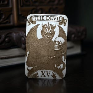 Könnte beinhalten: Eine rechteckige Tarotkarte mit einem gravierten Bild eines gehörnten Teufels mit Fledermausflügeln, der einen Schädel und eine Waffe hält. Die Karte ist oben mit "THE DEVIL" und unten mit "XV" beschriftet, mit einem detaillierten, strukturierten Design in Brauntönen.