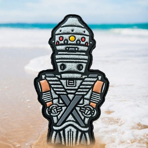 May include: A silver and black robot with a yellow and red eye patch, standing on a sandy beach with white foam waves.