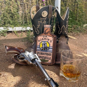 May include: A brown leather cowboy boot with a brown leather patch on the side. The patch features a cartoon image of a bottle with the text "Dr. Djarin's Splendiferous Sarsaparilla".