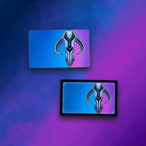 May include: Two versions of a Mandalorian symbol patch. The symbol is a silver, stylised, geometric design on a blue and pink background. The patch on the left is a sticker, while the patch on the right is sewn onto a black fabric.