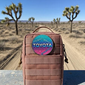 May include: A dusty rose-colored tactical pouch with a circular patch featuring a mountain landscape design and the word "TOYOTA". The pouch has a handle and multiple webbing straps. The background shows a desert landscape with Joshua trees.