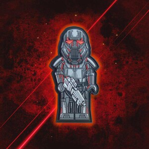 May include: A gray and red patch featuring a stylized figure in a helmet and armor, holding a blaster. The design has a geometric aesthetic with red accents and a black border. The background is a textured red.