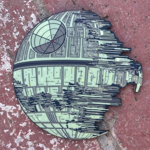 May include: A green and black Death Star pin with a damaged section. The pin is on a red brick surface.