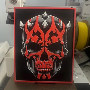 May include: A red and black skull with horns and a white outline. The skull is on a black background with a red border.