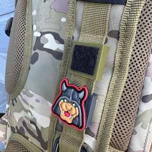 Pode incluir: Uma mochila de camuflagem verde com um remendo preto e vermelho de um monstro de desenho animado com chifres e um capacete. O remendo é preso a uma alça preta com fecho de velcro.