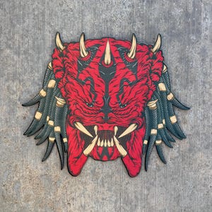 May include: A red and black Predator mask with sharp teeth and horns. The mask has a detailed design with black accents and green and tan dreadlocks. The artwork is set against a gray background.