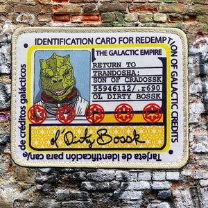 May include: A yellow and white identification card for the Galactic Empire with the text "IDENTIFICATION CARD FOR REDEMPTION OF GALACTIC CREDITS" and "THE GALACTIC EMPIRE". The card features a green alien with a toothy grin and the text "RETURN TO TRANDOSHA: SON OF CRADOSSK 55946112/.r690 OL DIRTY BOSSK". The card is signed "of Dirty Bosok".