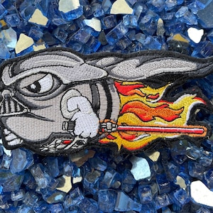 May include: A gray and black embroidered patch of a cartoon Darth Vader character with a red lightsaber, flying through the air with flames behind him.