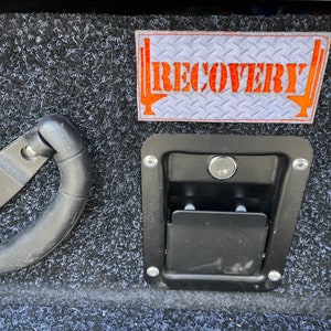 May include: A black truck bed with a grey carpet liner. A white patch with the word "RECOVERY" in orange letters is attached to the liner. A black handle and a black latch with a keyhole are also visible.