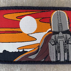 May include: A black and gray embroidered patch featuring a stylized silhouette of a figure wearing a helmet and a cape, standing in front of a sunset with a large white moon. The figure is facing away from the viewer.