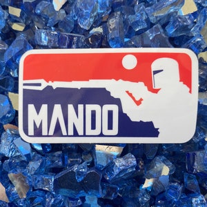 May include: A rectangular sticker with a red and blue design featuring a white silhouette of a character holding a weapon, with the word "MANDO" in white block letters. The sticker is set against a background of blue glass.