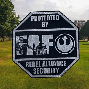 May include: Black and silver octagonal sign with the text "PROTECTED BY FAF REBEL ALLIANCE SECURITY". The sign features illustrations of C-3PO, R2-D2, and the Rebel Alliance symbol. The sign is placed on a green lawn.
