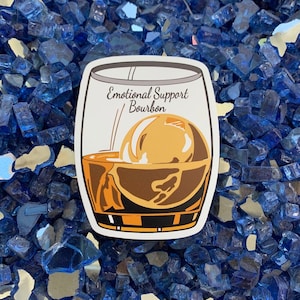 May include: A white sticker with a drawing of a glass of brown bourbon with an ice cube. The text on the sticker reads "Emotional Support Bourbon".