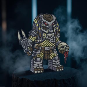 May include: Embroidered patch featuring a stylized Predator character. The patch showcases the Predator in a squat, cartoonish form, holding a skull with a red tongue. The design incorporates shades of gray, yellow, and brown.