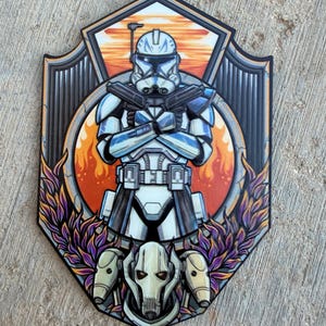 May include: A decorative wall plaque featuring a stylized illustration of a white and blue clone trooper with crossed blasters standing above a mechanical cyborg character. The design includes a circular orange background with flames and purple foliage.