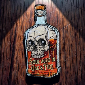 Può includere: Toppa ricamata a forma di bottiglia di liquore, con un disegno a teschio. La bottiglia contiene un liquido marrone-rossastro. Il testo recita "Soul Aged in Oak & Fire". La toppa ha un contorno azzurro chiaro e un tappo di sughero.