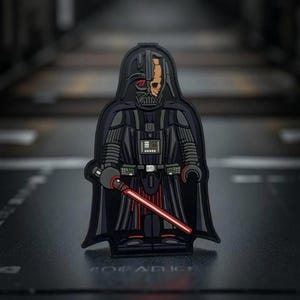 May include: A black and gray Darth Vader patch with a red lightsaber. The patch features a detailed design of the character, including his helmet and exposed face. The lightsaber is angled across the front.