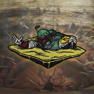 May include: An embroidered patch featuring a green and brown Star Wars Boba Fett character in a yellow sand dune. The patch is surrounded by a black border.