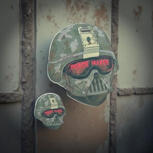 Op de afbeelding: Twee stickers met een helmdesign met een camouflagepatroon en de tekst "PEACE MAKER" in het rood. Het helmdesign is een mix van een militaire helm en de helm van een personage uit een sciencefictionfilm. Het woord "SKYWALKER" is ook zichtbaar.