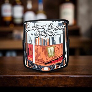 May include: A patch featuring a detailed illustration of a bourbon glass, filled with amber liquid. The patch has a black border and the text "Emotional Support Bourbon" in white script. The design is set against a blurred bar background.