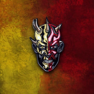 Pode incluir: Um remendo bordado que representa o rosto de um demônio com chifres. O demônio tem uma expressão feroz com dentes afiados e um queixo pontudo, metade do rosto é vermelho e a outra metade é amarela.