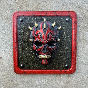 May include: A square wall plaque featuring a three-dimensional sculpted skull with red and black markings, gold-colored teeth, and small gold horns. The skull is mounted on a dark, textured square panel with a distressed red border and four rivets.