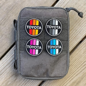 May include: A gray pouch with four circular patches featuring the "TOYOTA" logo in various color schemes: orange, red, yellow; gray and white; pink and white; and blue and white. The patches are black with white text.