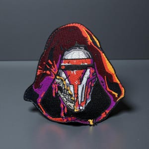 Może przedstawiać: Haftowana naszywka przedstawiająca postać w kapturze. Projekt zawiera hełm z czerwonymi, białymi i czarnymi detalami na czarnym tle. Kaptur jest w kolorze głębokiego bordowego z pomarańczowymi i fioletowymi akcentami.