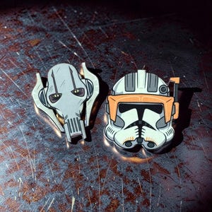 May include: Two enamel pins featuring characters from a science fiction franchise. One pin depicts a gray, skeletal face with yellow eyes. The other pin shows a white and orange helmet with black details. Both pins are on a textured, dark surface.