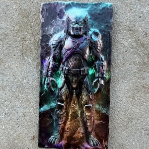May include: A rectangular wall plaque featuring a three-dimensional relief of the Predator character. The figure is rendered in dark metallic tones with vibrant, glowing green and purple highlights across the textured, rocky background surface.