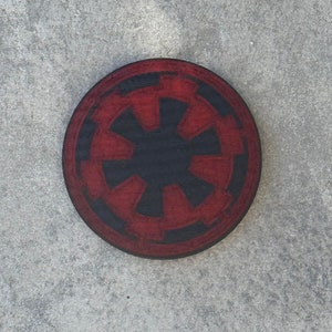 May include: A round, wooden coaster featuring a red and black Imperial symbol. The coaster has a dark red outer ring and a black center with a starburst design. The coaster is likely for use with beverages.