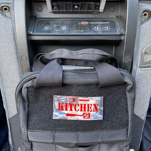 May include: A black tactical backpack with a red patch that says "Kitchen" on it. The backpack is sitting in front of a black and silver ARB Fridge Freezer.