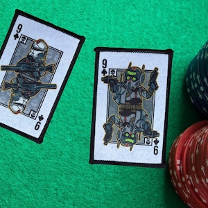 Może przedstawiać: Dwie karty do gry z ilustracją żołnierza, oznaczone numerem dziewięć i kolorem trefl. Karty leżą na zielonej powierzchni, w tle stosy żetonów do pokera.