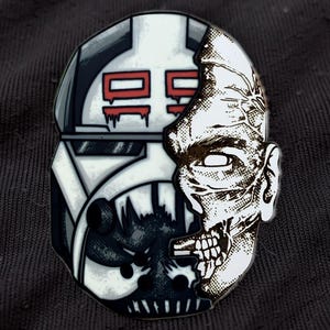 Puede incluir: Un pin de esmalte metálico con un diseño dividido. El lado izquierdo muestra un casco blanco y gris con marcas rojas y el número 99. El lado derecho muestra una ilustración detallada en blanco y negro de un rostro humano.