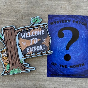 Pode incluir: Um remendo bordado azul e marrom com um Ewok de desenho animado se escondendo atrás de uma árvore com uma placa que diz "Bem-vindo a Endor!" O remendo faz parte de um programa de remendo misterioso do mês.