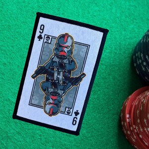 May include: A rectangular patch featuring a playing card design with a cartoon soldier illustration. The card is the nine of clubs, with the number 9 at the top left and 6 at the bottom right. The patch is on a green surface with poker chips.
