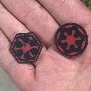 May include: Two black and red Star Wars themed tokens. One is a hexagon with a six-pointed star design, and the other is a circle with a similar starburst pattern. The tokens are held in a hand.