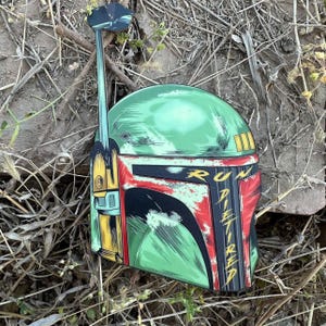 May include: A green, red, and yellow helmet with the text "RUN DIETIRED". The helmet has a long antenna and is lying on a bed of dry grass and small rocks. The helmet is a Star Wars themed item.