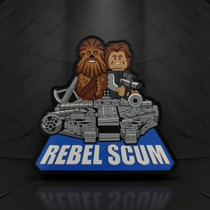 May include: A patch featuring Chewbacca and Han Solo, with the Millennium Falcon, in a Lego-like style. The patch is gray, brown, and blue, with the words "REBEL SCUM" in white on a blue background.