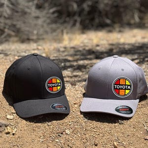 Pode incluir: Duas bonés de beisebol Flexfit, uma preta e outra cinza, cada uma com um logotipo circular da Toyota em vermelho, amarelo e laranja. Os bonés são exibidos em uma superfície de terra, mostrando seu design e estilo.