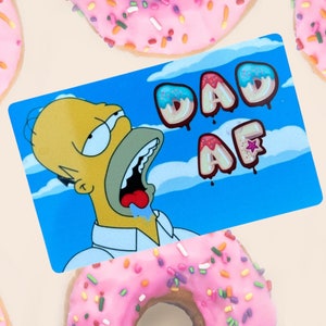 May include: A blue background with white clouds and the words "DAD AF" spelled out in pink and blue donut letters. A cartoon image of Homer Simpson with his mouth open and drool coming out is in the foreground.