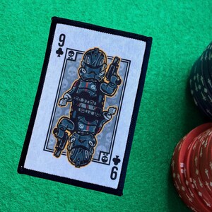 May include: A playing card patch featuring a soldier-like figure with a gun, set against a white background with the numbers 9 and 6. The patch is on a green surface, with stacks of poker chips in the background.